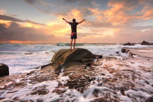 Teen boy stands on a rock among turbulent ocean seas and fast flowing water at sunrise. Worship praise zest adenture solitude finding peace among lifes turbulent times. Overcoming adversity. Motion in water
